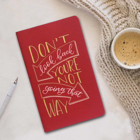 hand painted journal cover that says don't look back you're not going that way in block lettering and modern calligraphy with arrow doodles shown on the cranberry journal cover and gold and pink paint shown laying next to a white pen, cozy knitted blanket and cup of coffee