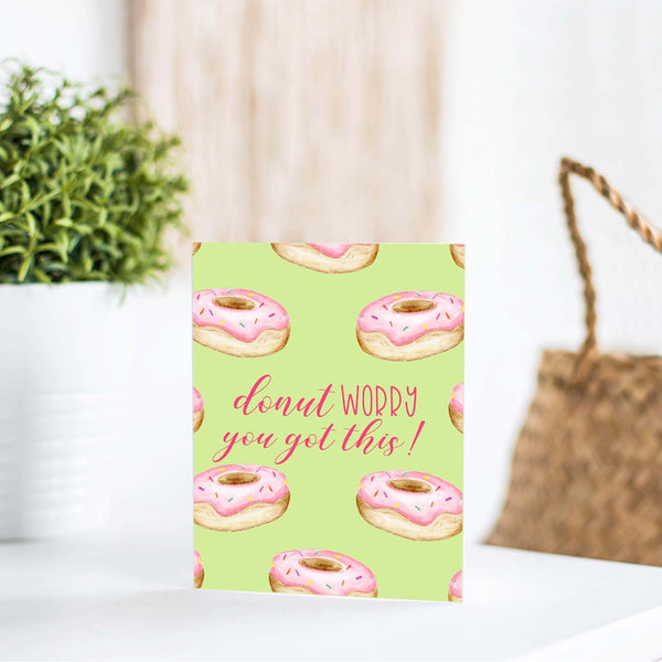 watercolor pink frosted donuts with sprinkles on a greeting card that says donut worry you got this with a white A2 envelope  shown standing on a white table with a plant and a handbag