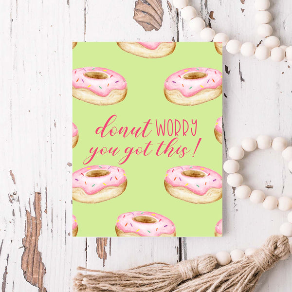 watercolor pink frosted donuts with sprinkles on a greeting card that says donut worry you got this with a white A2 envelope shown laying on a rustic white wood table with white wood bead garland