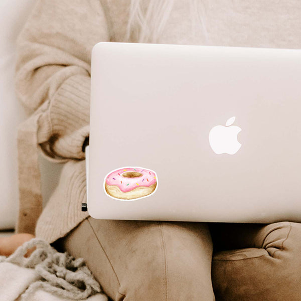 3" vinyl watercolor sticker of a a round donut with pink frosting and rainbow sprinkles shown on a MacBook cover sitting open on a woman's lap