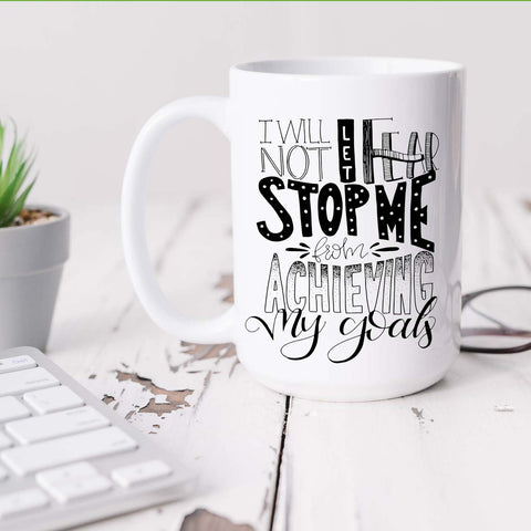 15oz white ceramic mug with hand lettered illustrated design that says I will not let fear stop me from achieving my goals shown sitting on a white desk with plant, keyboard adn glasses