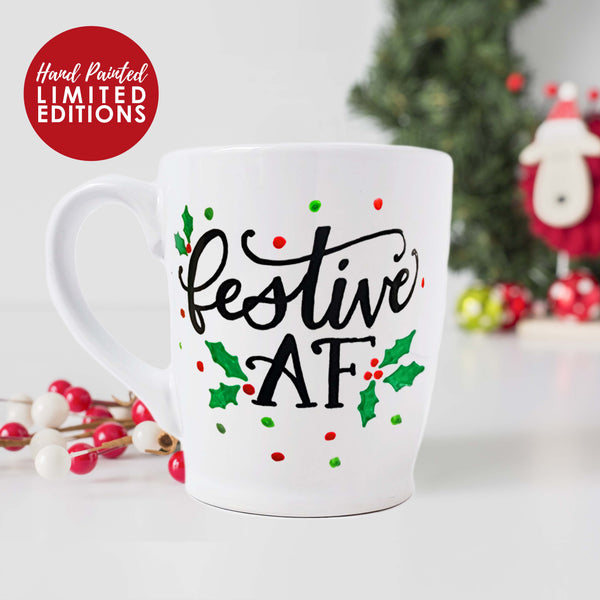 Hand painted white ceramic mug that says festive AF in black hand lettering surrounded by holly leaves, berries and green and red polka dots