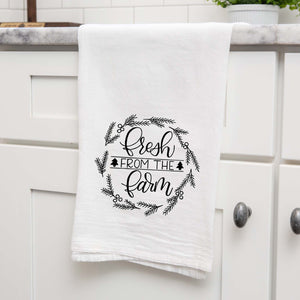White Floursack Kitchen towel with black hand illustrated design that says fresh from the farm with twigs and berries in a wreath shape shown hanging from the counter in a kitchen