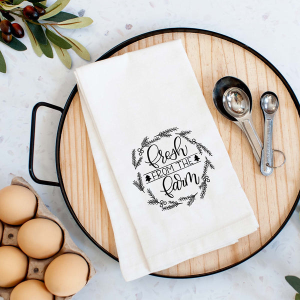 White Floursack Kitchen towel with black hand illustrated design that says fresh from the farm with twigs and berries in a wreath shape shown folded on a tray with measuring spoons and fresh eggs in a kitchen