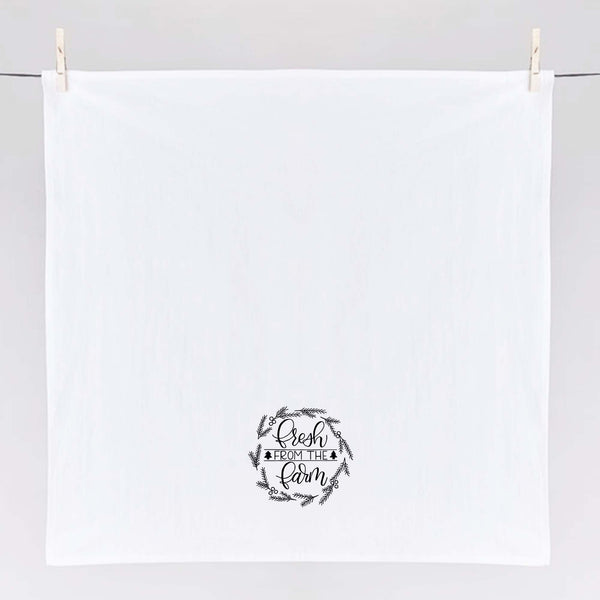 White Floursack Kitchen towel with black hand illustrated design that says fresh from the farm with twigs and berries in a wreath shape shown hanging unfolded from clothes pins