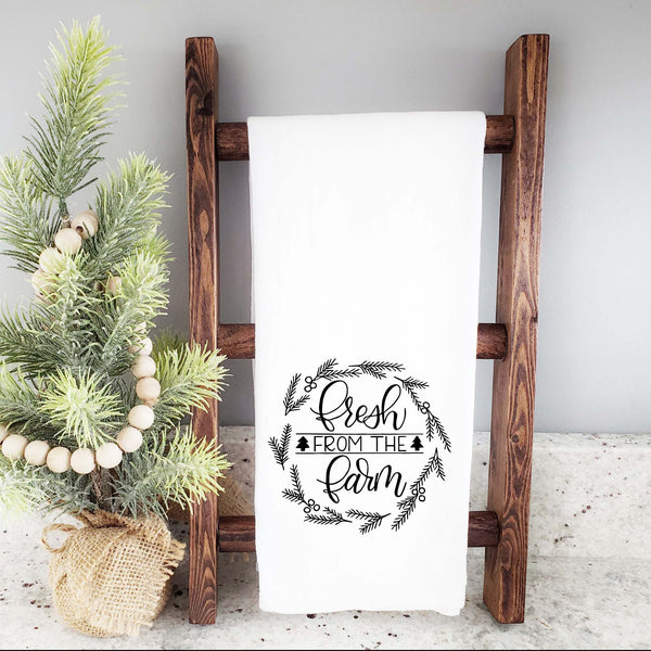 White Floursack Kitchen towel with black hand illustrated design that says fresh from the farm with twigs and berries in a wreath shape shown hanging from a wooden display ladder next to a mini Christmas tree
