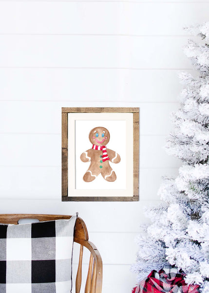 Watercolor gingerbread man painting with a red and white striped scarf shown in a wood frame hanging on a shiplap wall next to a white flocked tree and rocking chair.