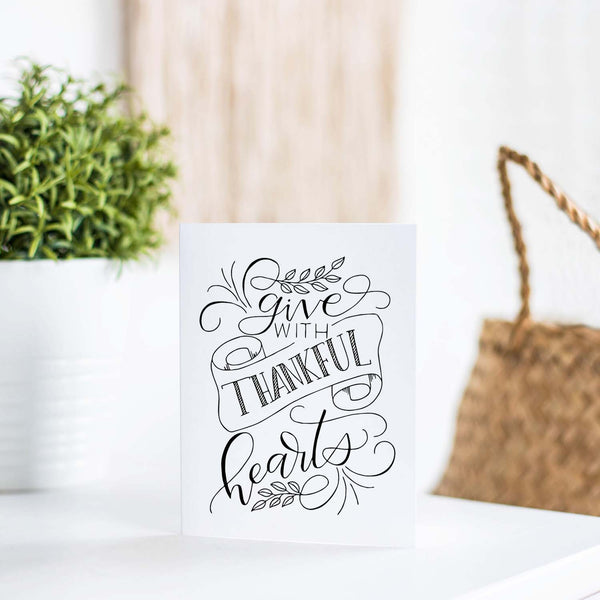 Give with thankful hearts hand lettered greeting card design in black on a folded white card with A2 envelope shown on a white table with plant and handbag