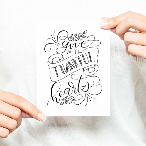 Give with thankful hearts hand lettered greeting card design in black on a folded white card with A2 envelope shown with a woman in a white sweater holding card