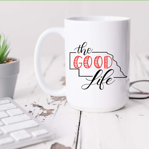 15oz white ceramic mug with hand lettered illustrated design that says The Good Life with the outline of the state of Nebraska shown sitting on a white office desk