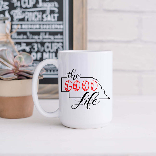15oz white ceramic mug with hand lettered illustrated design that says The Good Life with the outline of the state of Nebraska shown sitting in a kitchen