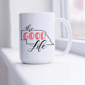 15oz white ceramic mug with hand lettered illustrated design that says The Good Life with the outline of the state of Nebraska shown sitting in a sunny window