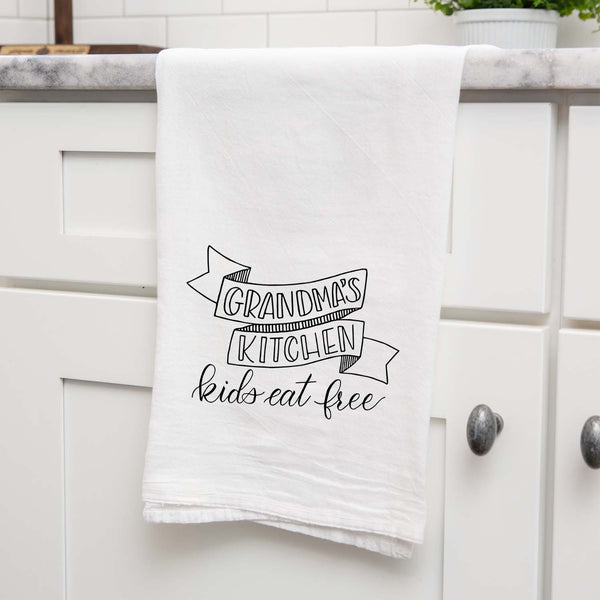 White floursack kitchen towel with black hand lettered illustration that says grandma's kitchen kids eat free shown hanging folded from a countertop in a modern kitchen