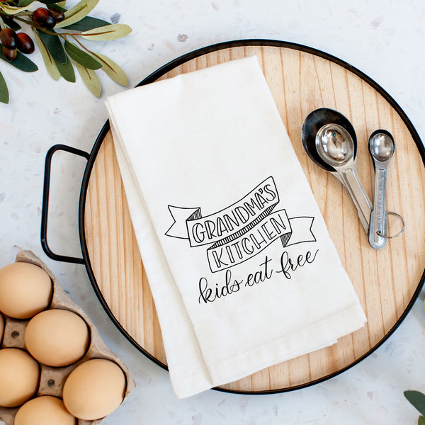 White floursack kitchen towel with black hand lettered illustration that says grandma's kitchen kids eat free shown folded on a serving tray with a set of measuring spoons and fresh eggs
