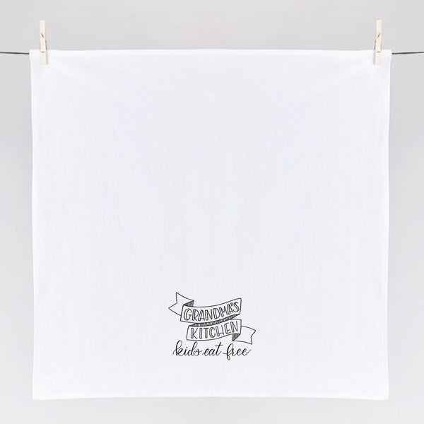 White floursack kitchen towel with black hand lettered illustration that says grandma's kitchen kids eat free shown hanging from clothes pins