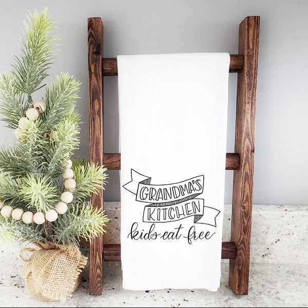 White floursack kitchen towel with black hand lettered illustration that says grandma's kitchen kids eat free shown hanging folded from a wooden display ladder with a mini Christmas tree