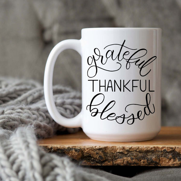 15oz white ceramic mug with hand lettered illustrated design that says Grateful Thankful Blessed shown sitting on a wood tray with a grey knit blanket