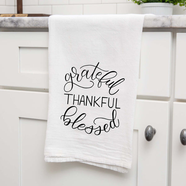 White floursack towel with black hand illustrated design that says grateful thankful blessed shown folded and hanging from a countertop in a modern kitchen