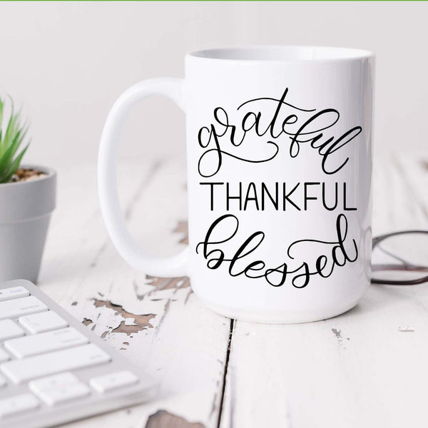 15oz white ceramic mug with hand lettered illustrated design that says Grateful Thankful Blessed shown sitting on a white office desk