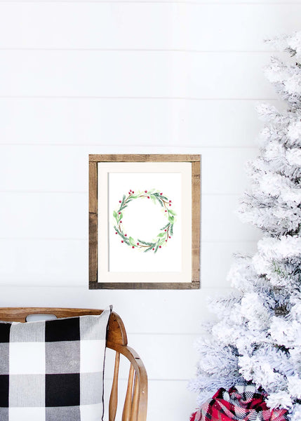 Watercolor painted holiday wreath with greens, branches and red berries shown in a wooden frame hanging on a shiplap wall with a white flocked christmas tree and rocking chair
