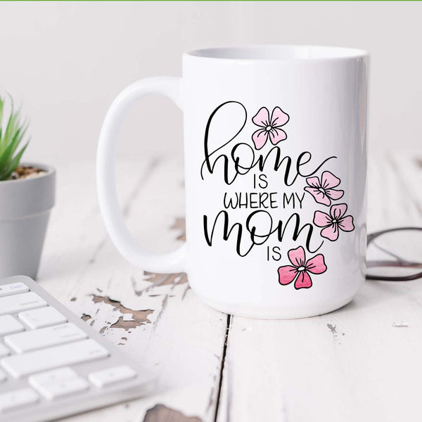 15oz white ceramic mug with hand lettered illustrated design that says home is where my mom is with pink flower illustrations shown on a white office desk