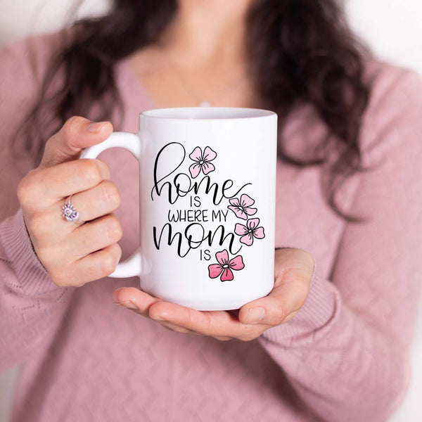 15oz white ceramic mug with hand lettered illustrated design that says home is where my mom is with pink flower illustrations shown with a woman holding the mug