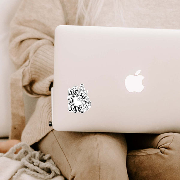 3" black and white illustrated hand lettered vinyl sticker says I need a drink with an illustrated hanging pothos plant shown adhered to a MacBook laptop cover sitting open on a woman's lap
