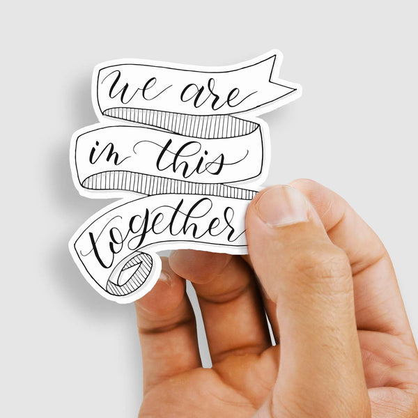 3" hand lettered, illustrated, black and white vinyl sticker that says we are in this together with a ribbon banner illustration shown with a woman's hand holding the sticker
