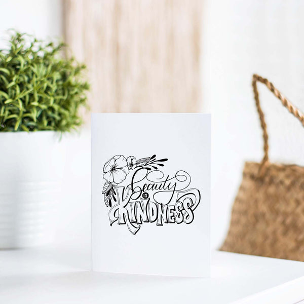 Beauty is kindness hand lettered and illustrated black and white greeting card on a folded white card with an A2 envelope shown on a white table with a plant and handbag