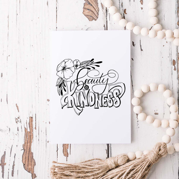 Beauty is kindness hand lettered and illustrated black and white greeting card on a folded white card with an A2 envelope shown on a white rustic table with white wooden bead garland