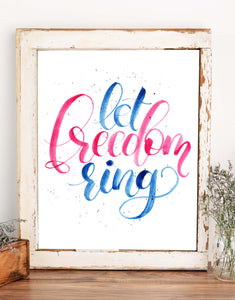Hand painted watercolor hand lettering that says let freedom ring in patriotic red and blue shown in a rustic frame on a shelf