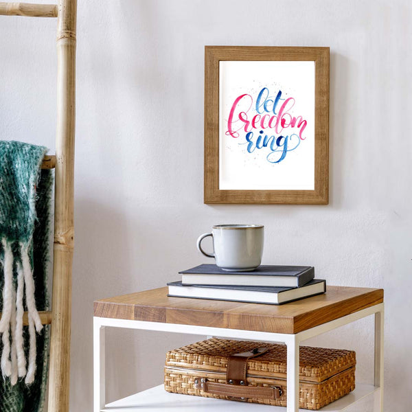 Hand painted watercolor hand lettering that says let freedom ring in patriotic red and blue shown hanging in a living room by a table with books and coffee mug