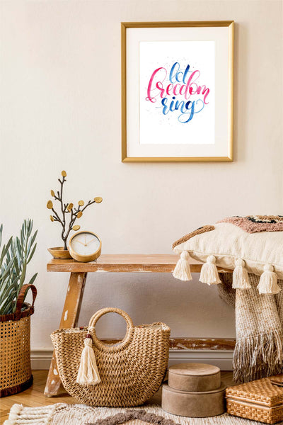 Hand painted watercolor hand lettering that says let freedom ring in patriotic red and blue shown hanging in a living room with a bench, throw pillows and other decor