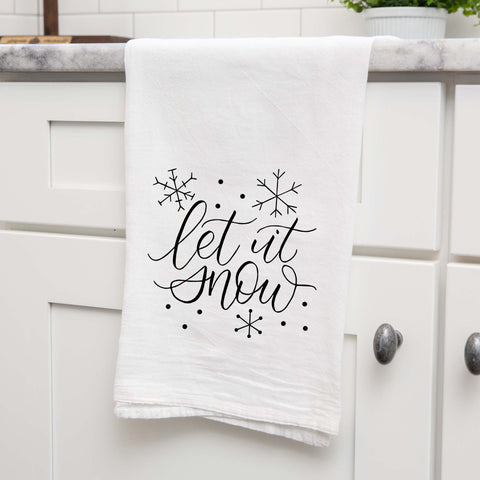 White Floursack Kitchen Towel with black hand lettered illustration that says let it snow with snowflake doodles shown hanging from a countertop in a modern kitchen