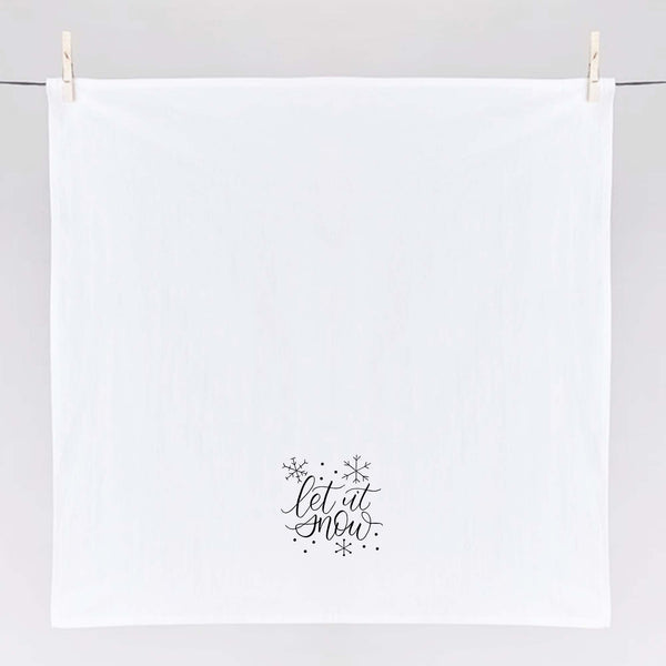 White Floursack Kitchen Towel with black hand lettered illustration that says let it snow with snowflake doodles shown hanging unfolded from clothes pins