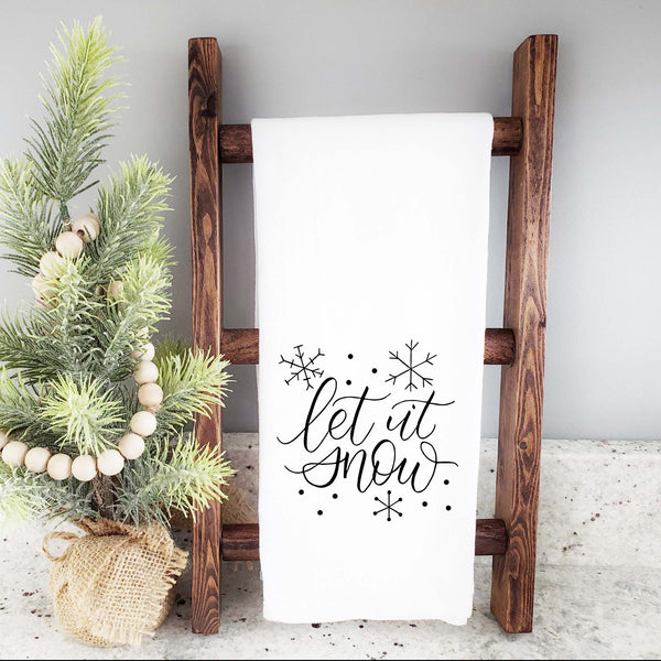White Floursack Kitchen Towel with black hand lettered illustration that says let it snow with snowflake doodles shown hanging folded from a wooden display ladder with a mini Christmas Tree