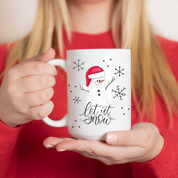 15oz white ceramic mug with watercolor snowman with a red santa hat, snowflake doodles and says let it snow in calligraphy shown with a woman in a red holiday sweater holding the mug