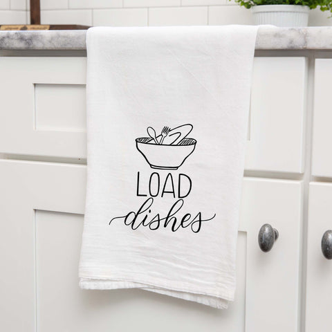 White floursack towel with black hand lettered illustrated design that says Load dishes with pile of dirty dishes doodles shown folded and hanging from a countertop in a modern kitchen