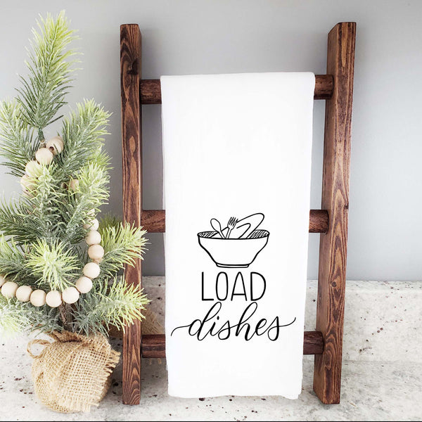 White floursack towel with black hand lettered illustrated design that says Load dishes with pile of dirty dishes doodles shown folded and hanging from a wooden display ladder with a mini Christmas tree