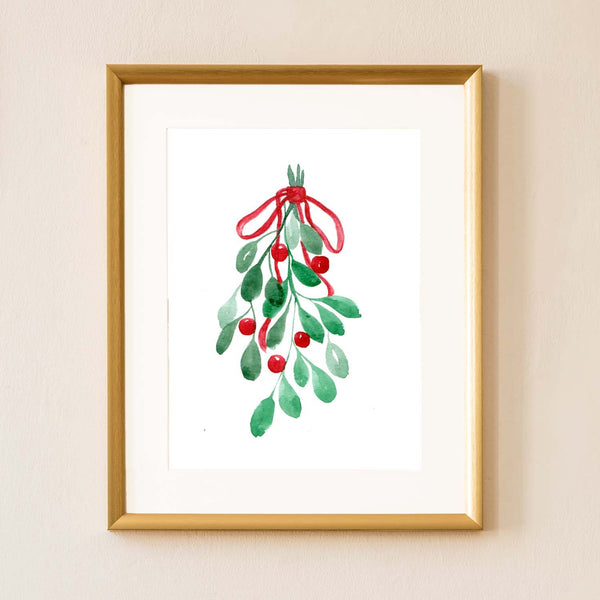 Watercolor painting of mistletoe tied with a red ribbon shown hanging on a wall in a gold frame