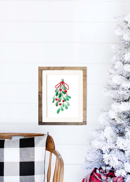 Watercolor painting of mistletoe tied with a red ribbon shown in a wood frame hanging on a shiplap wall next to a white flocked tree and rocking chair
