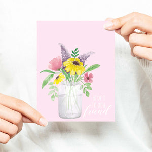 watercolor mason jar filled with colorful wild flowers friendship greeting card that says here's to you, friend with a white A2 envelope shown with a woman in a white sweater holding card
