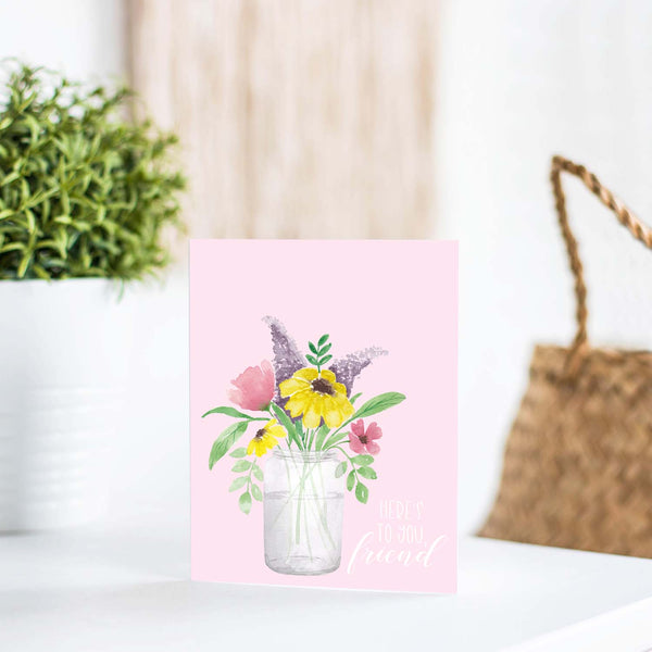 watercolor mason jar filled with colorful wild flowers friendship greeting card that says here's to you, friend with a white A2 envelope shown standing on a white table with a plant and handbag
