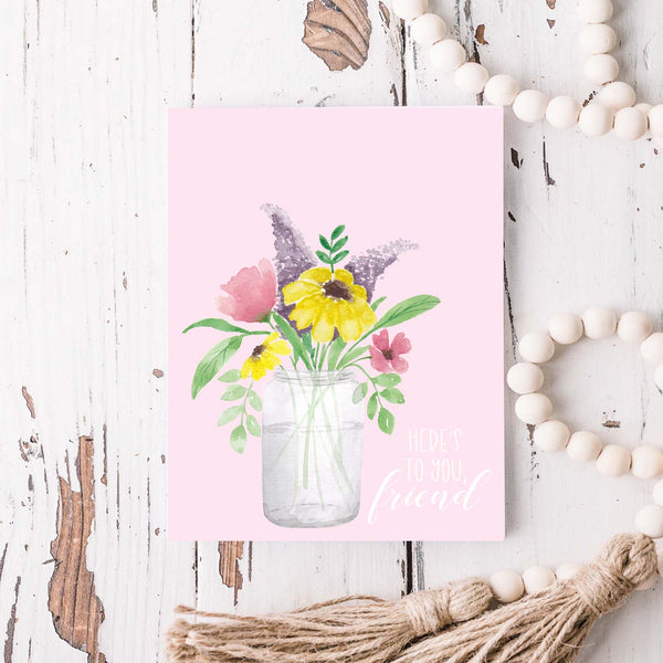 watercolor mason jar filled with colorful wild flowers friendship greeting card that says here's to you, friend with a white A2 envelope shown laying on a rustic white wood table with a white wooden bead garland