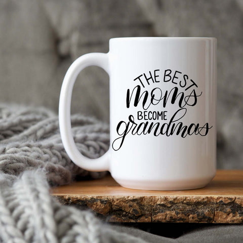 15oz white ceramic mug with hand lettered illustrated design that says the best moms become grandmas shown sitting on a wood tray with a grey knit blanket
