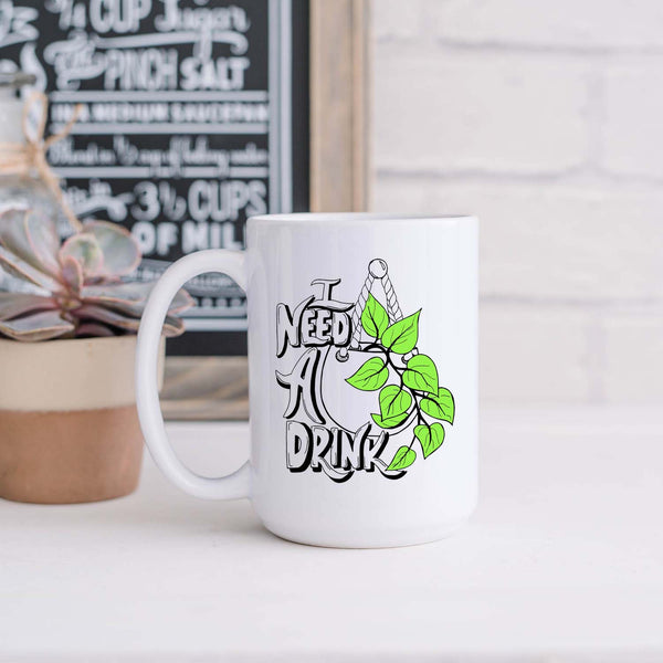 15oz white ceramic mug with hand lettered illustrated design that says I need a drink with an illustration of a pothos plant with green leaves in a hanging pot shown sitting in a kitchen