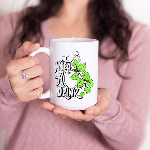 15oz white ceramic mug with hand lettered illustrated design that says I need a drink with an illustration of a pothos plant with green leaves in a hanging pot shown with a woman holding the mug