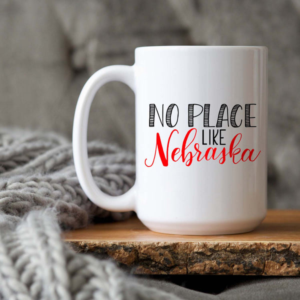 15oz white ceramic mug with hand lettered illustrated design that says No Place Like Nebraska shown on a wood tray with a grey knit blanket