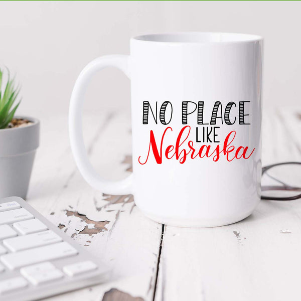 15oz white ceramic mug with hand lettered illustrated design that says No Place Like Nebraska shown sitting on a white office desk
