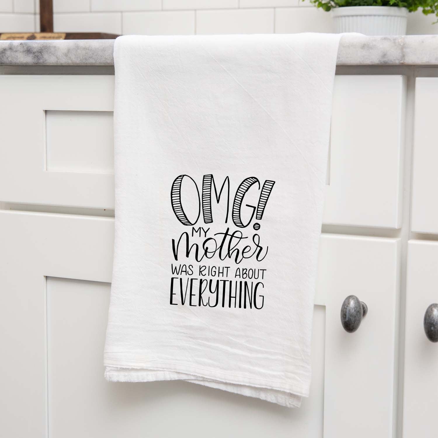 White floursack kitchen towel with black hand lettered illustrated design that says OMG! My mother was right about everything shown folded and hanging from a countertop in a modern kitchen